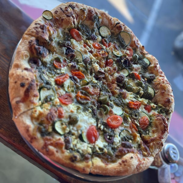 A freshly baked pizza with pesto, tomatoes, zucchini, olives, and feta cheese on a wooden table.